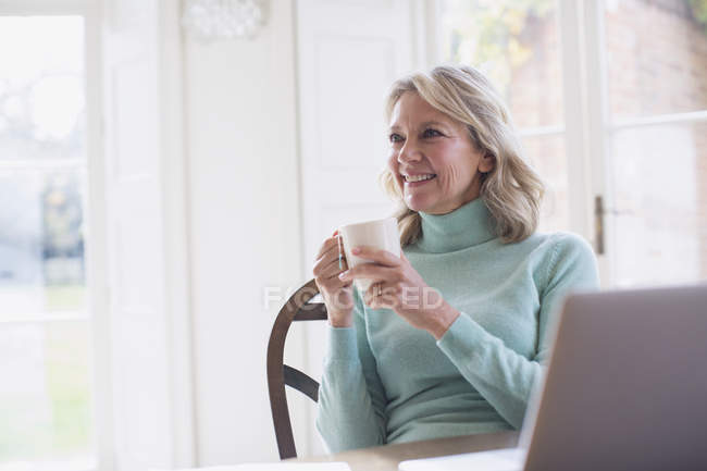 Улыбающаяся взрослая женщина-фрилансер пьет чай и работает дома за ноутбуком — стоковое фото