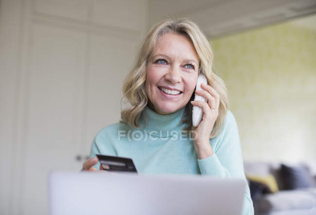 Улыбающаяся взрослая женщина с кредитной картой разговаривает по телефону за ноутбуком — стоковое фото
