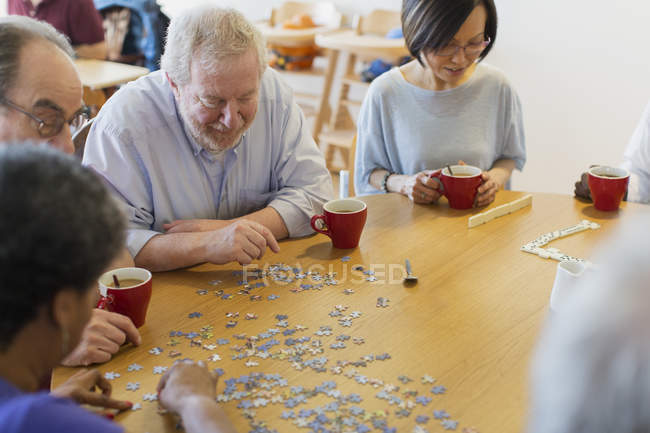 Senior friends assembling jigsaw puzzle and drinking tea at table in community center — Stock Photo