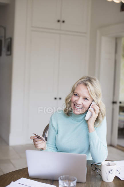 Усміхнена зріла жінка з кредитною карткою розмовляє по телефону в ноутбуці — стокове фото