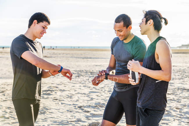 Coureurs masculins vérifiant smart watch fitness trackers sur la plage ensoleillée — Photo de stock