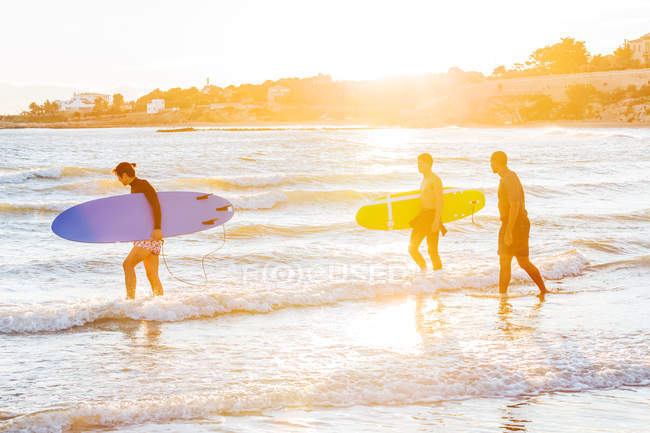 Surfistas masculinos transportando pranchas de surf para o oceano na praia ensolarada — Fotografia de Stock