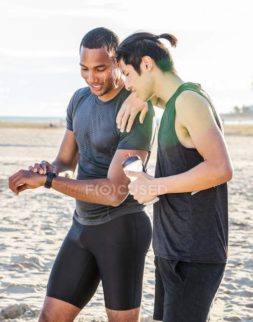 Corredores masculinos descansando, comprobando rastreador de fitness reloj inteligente en la playa soleada - foto de stock