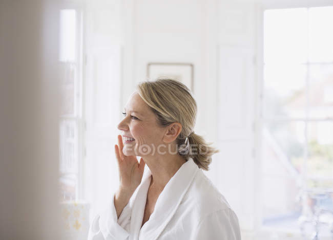 Улыбающаяся зрелая женщина наносит увлажняющий крем на лицо в ванной комнате — стоковое фото