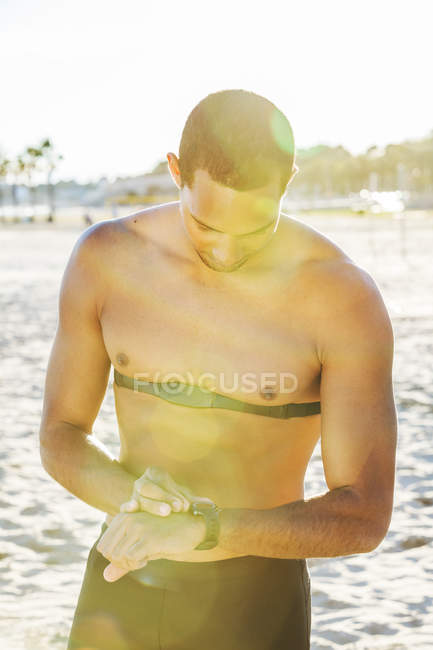 Corredor masculino con rastreador de fitness comprobando reloj de pulsera en la playa soleada - foto de stock