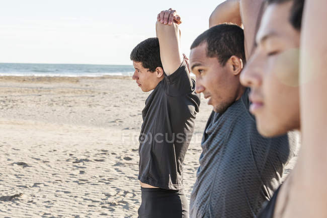 Corredores masculinos estirando los brazos en la playa soleada - foto de stock