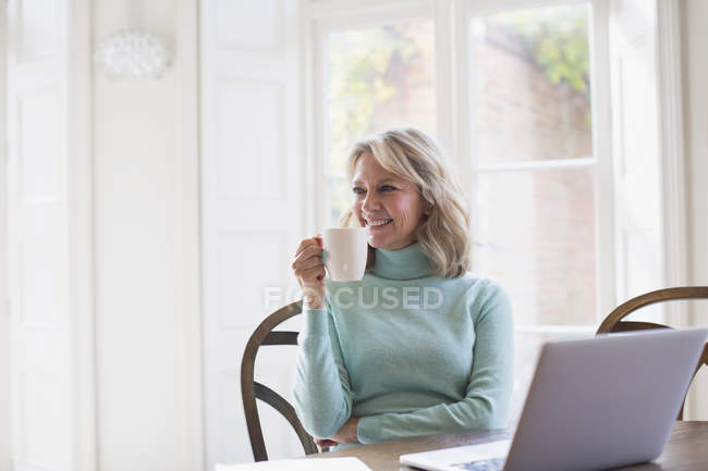 Sonriendo madura freelancer bebiendo té y trabajando en el ordenador portátil en casa - foto de stock