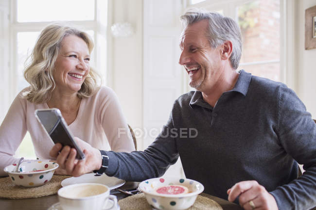 Laughing mature couple using smart phone and eating breakfast — Stock Photo