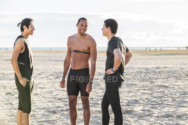 Corredores masculinos descansando y hablando en la playa soleada - foto de stock