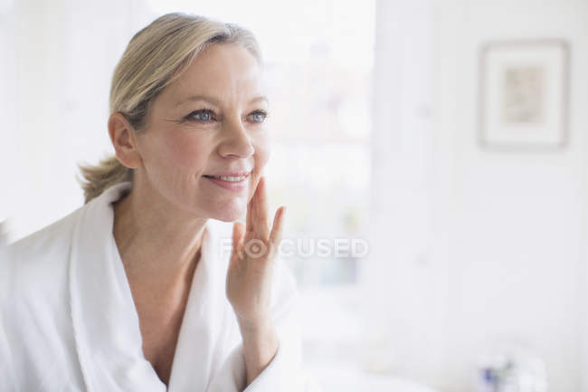 Улыбающаяся зрелая женщина, накладывающая увлажняющий крем на лицо перед зеркалом в ванной — стоковое фото