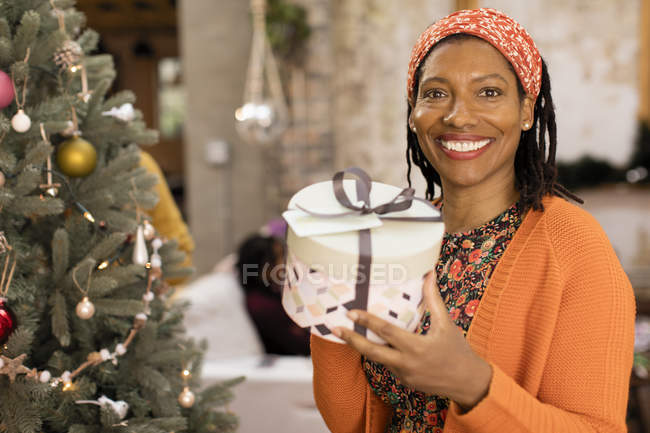 Retrato sonriente, mujer confiada sosteniendo regalo al lado del árbol de Navidad - foto de stock