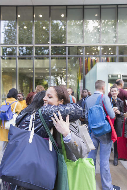 Estudiantes universitarias con bolsas abrazándose fuera del edificio - foto de stock