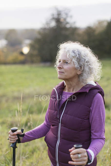 Thoughtful active senior woman hiking with poles in rural field — Stock Photo