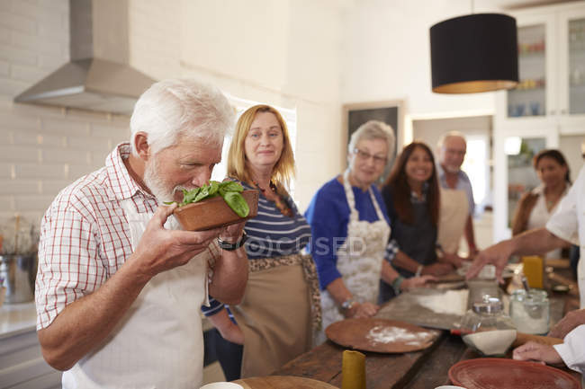 Amigos mayores activos tomando clases de cocina, oliendo albahaca fresca - foto de stock