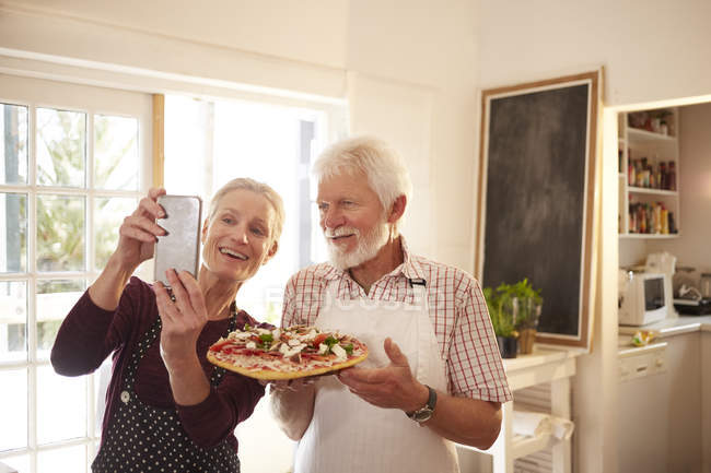 Sonriente y segura pareja de ancianos tomando selfie con pizza en la clase de cocina - foto de stock