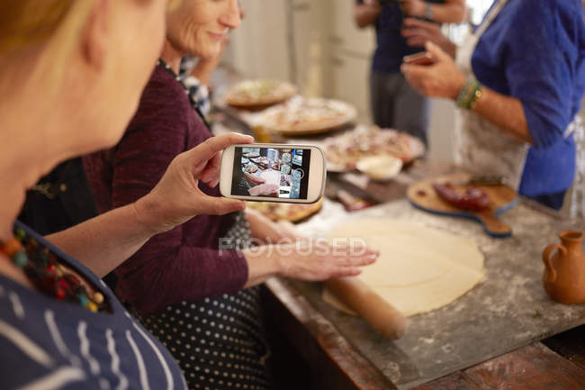 Mujer con cámara de teléfono fotografiando amigo haciendo masa de pizza en clase de cocina - foto de stock