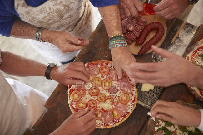 Overhead view senior friends adding tomatoes and meat to fresh pizza — Stock Photo