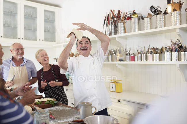 Amigos mayores viendo al juguetón chef lanzando masa de pizza en clase de cocina - foto de stock