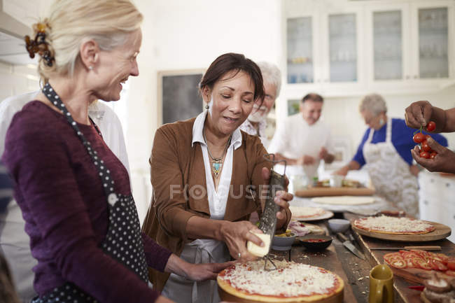 Mujeres mayores amigas rallando queso sobre pizza en clase de cocina - foto de stock