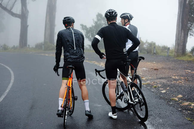 Amigos ciclistas masculinos tomando un descanso, descansando en el camino mojado - foto de stock