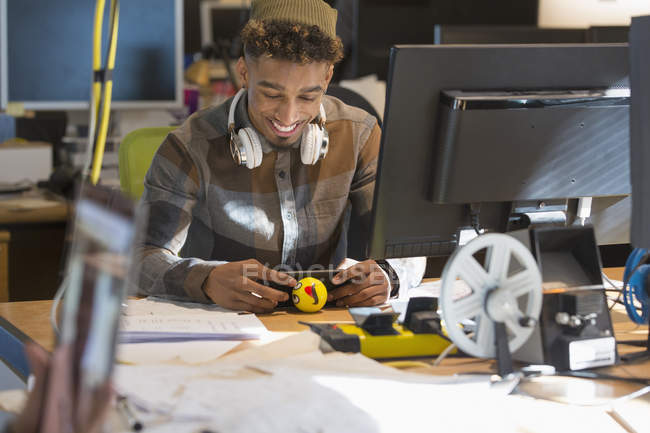 Улыбающийся креативный бизнесмен с наушниками, использующий смартфон на компьютере в офисе — стоковое фото
