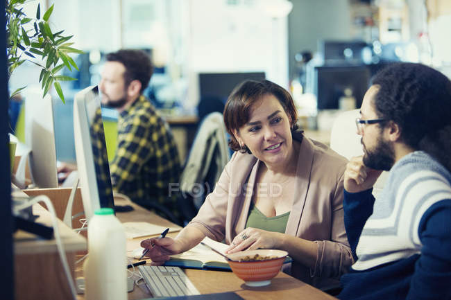 Creative business people eating cereal, meeting at computer in open plan office — Stock Photo