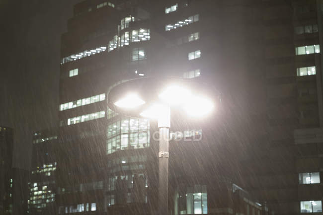 Rain falling around streetlamp below urban highrise buildings at night — Stock Photo