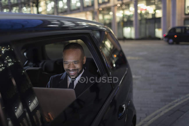 Empresário sorrindo usando laptop no táxi crowdsourced à noite — Fotografia de Stock