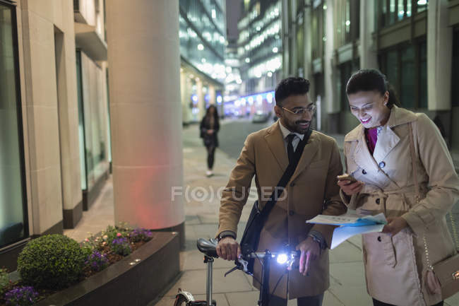 Business people with smart phone and bicycle reviewing paperwork on urban street at night — Stock Photo