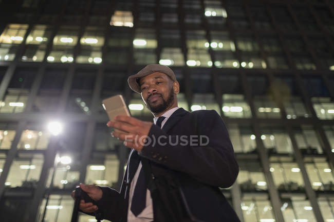 Empresário com telefone inteligente em pé abaixo do arranha-céus urbano à noite — Fotografia de Stock