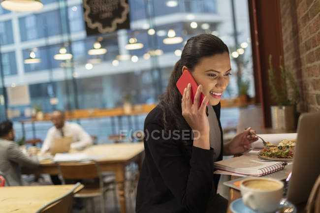 Улыбающаяся деловая женщина разговаривает на смартфоне и работает за ноутбуком в кафе — стоковое фото