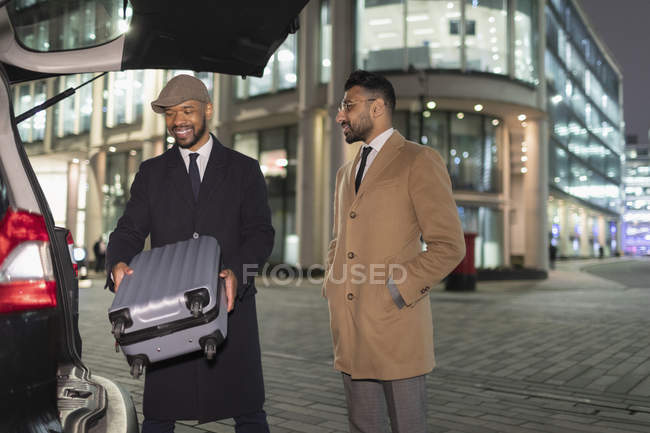 Des hommes d'affaires chargent une valise dans une voiture au coin d'une rue urbaine la nuit — Photo de stock