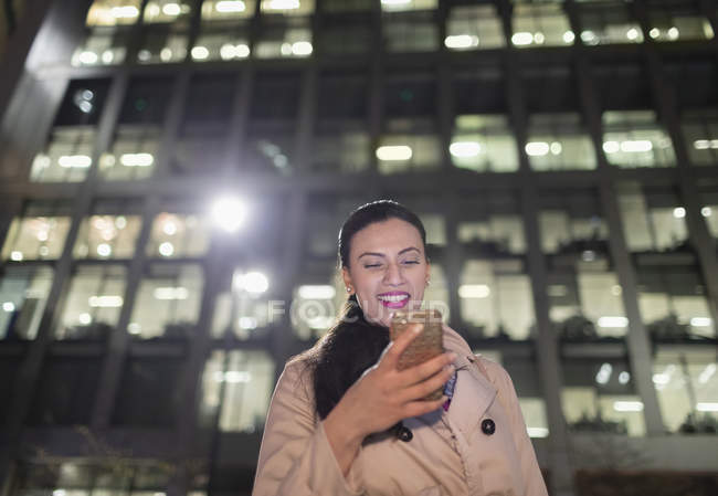 Smiling businesswoman using smart phone below urban highrise at night — Stock Photo
