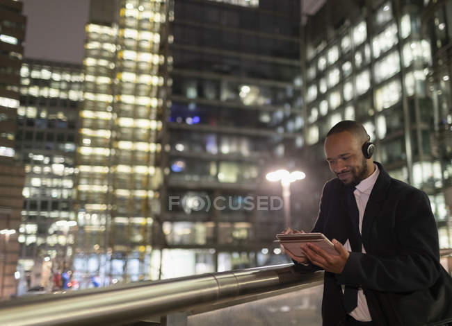 Uomo d'affari con cuffie utilizzando tablet digitale sul ponte pedonale urbano di notte — Foto stock