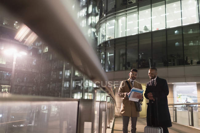 Businessmen with suitcase discussing paperwork on urban pedestrian bridge at night — Stock Photo