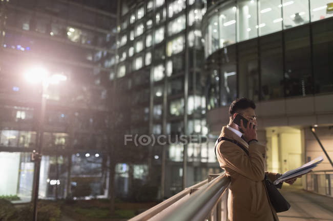Businessman talking on smart phone, reading paperwork on urban pedestrian bridge at night — Stock Photo