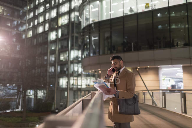 Businessman talking on smart phone and reading paperwork on urban pedestrian bridge at night — Stock Photo