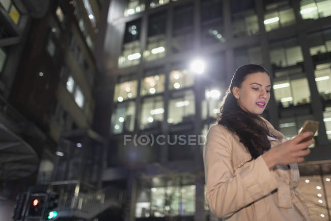 Businesswoman using smart phone below urban highrise at night — Stock Photo