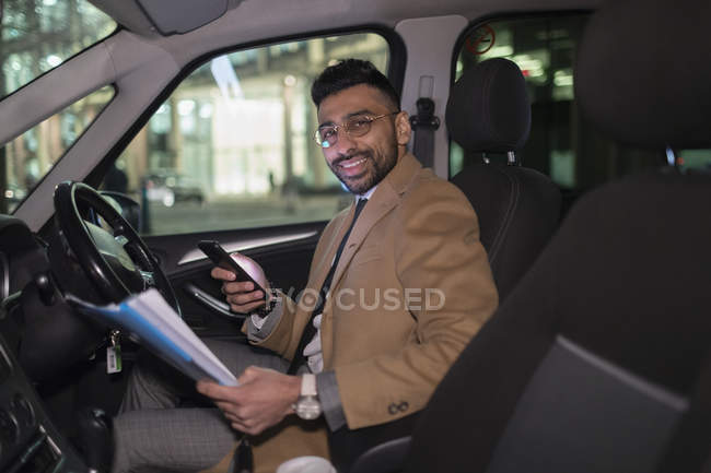 Retrato sorridente empresário com telefone inteligente leitura papelada no carro à noite — Fotografia de Stock