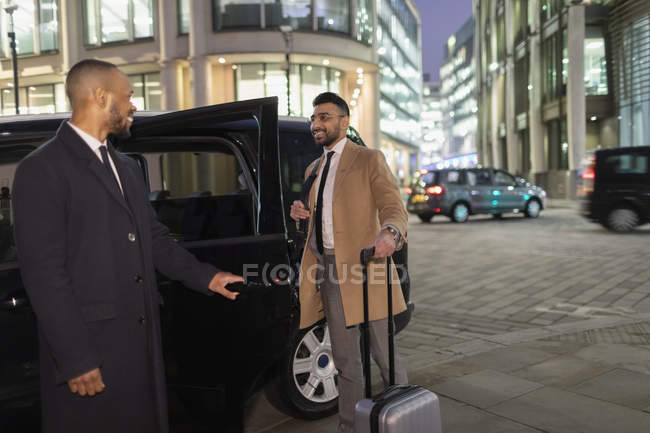 Motorista de abertura de porta de carro para homem de negócios com mala na rua urbana à noite — Fotografia de Stock