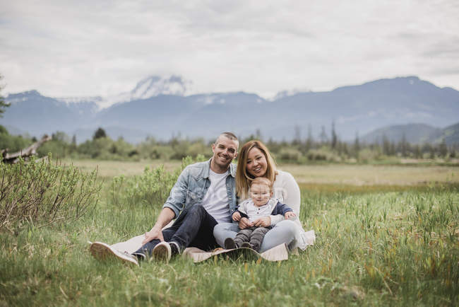 Ritratto genitori sorridenti e bambino seduto nel campo rurale con le montagne sullo sfondo — Foto stock
