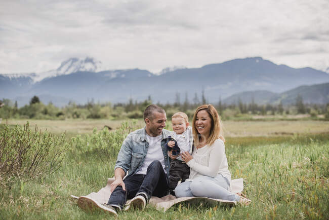 Parents et bébé fils assis dans un champ rural avec des montagnes en arrière-plan — Photo de stock