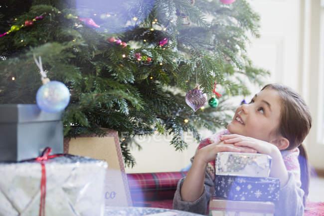 Curious girl with stack of gifts looking up at Christmas tree — Stock Photo