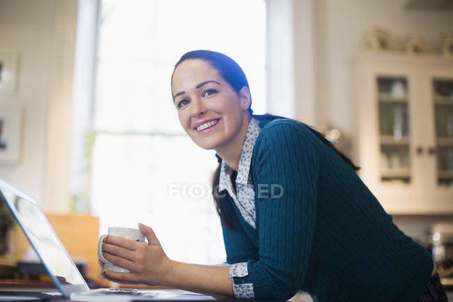 Retrato confiado, mujer sonriente beber café y trabajar en el ordenador portátil en la cocina - foto de stock