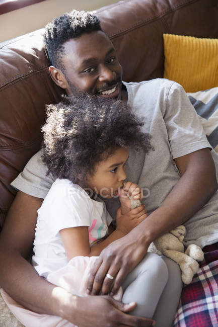Portrait affectionate father cuddling with toddler daughter sucking thumb on sofa — Stock Photo