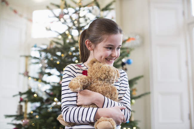 Happy girl hugging teddy bear in front of Christmas tree — Stock Photo