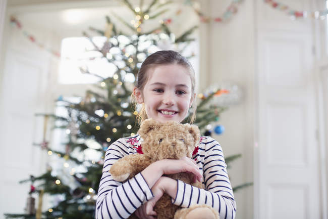 Ritratto sorridente, ragazza carina che tiene l'orsacchiotto davanti all'albero di Natale — Foto stock