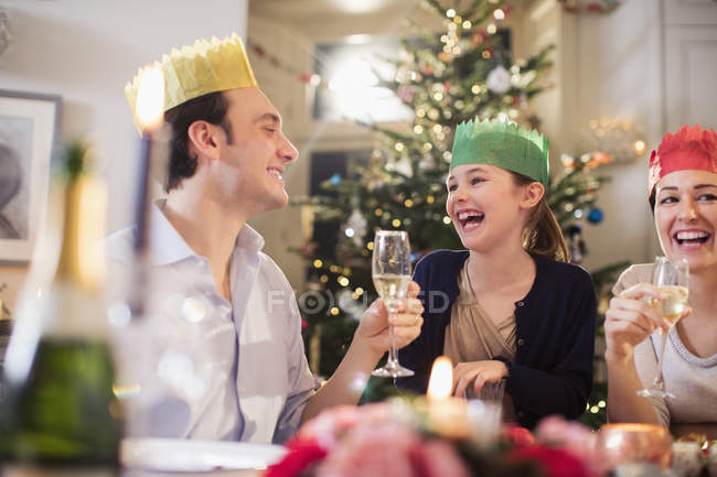 Familia feliz en coronas de papel disfrutando de la cena de Navidad, bebiendo champán y riendo - foto de stock