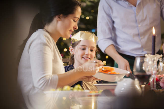 Mother serving carrots to daughter in paper crown at Christmas dinner — Stock Photo