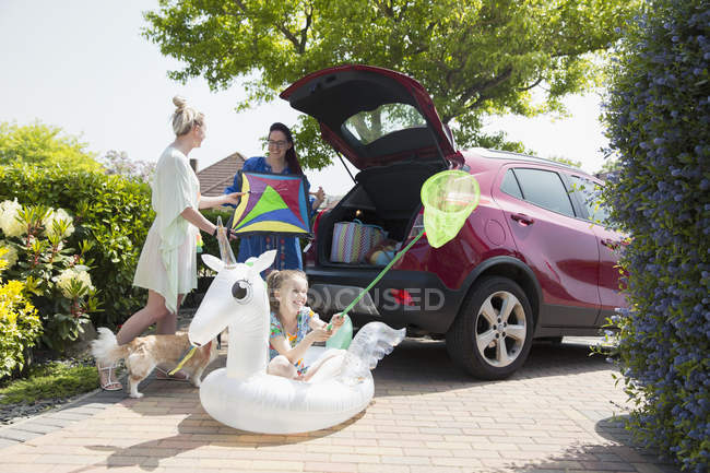 Lesbian couple and daughter loading kite and inflatable unicorn into car hatchback in sunny driveway — Stock Photo